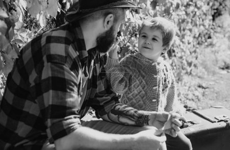 Foto de Joven padre hipster tatuado barbudo habla con su pequeño hijo sonriente. Padre e hijo se relajan en un banco después de caminar por el bosque. Feliz caminata familiar en el soleado día otoñal - Imagen libre de derechos