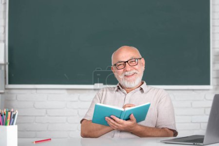 Foto de Hombre maestro maduro. Retrato de feliz tutor senior en gafas con libro en la pizarra en clase en la escuela secundaria o collage. Estudiante de edad en la universidad - Imagen libre de derechos