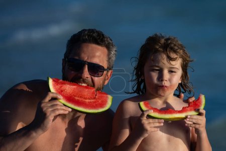 Téléchargez les photos : Père et fils mangent pastèque rouge juteuse sur la plage. Vacances d'été pour enfants, fruits de saison et baies. Été père et fils fils portrait extérieur. Visage enfant d'été, fruits d'été - en image libre de droit