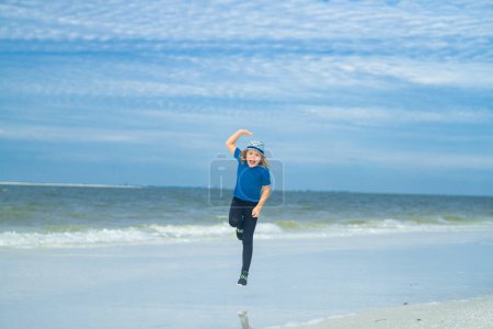 Foto de El chico corre en la playa de verano. Niño corriendo a lo largo del océano. El niño corre a lo largo de la orilla del mar en las vacaciones de verano. Feliz niño descansa en el mar. Concepto vacaciones de verano - Imagen libre de derechos