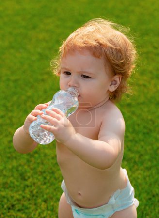 Foto de Bebé bebiendo agua. Primer plano del niño rubio bebiendo agua fresca y pura de la botella con un fondo de hierba verde borrosa - Imagen libre de derechos