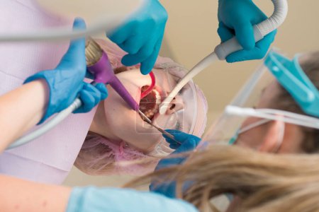 Téléchargez les photos : Portrait rapproché d'une patiente chez un dentiste de la clinique. Jeune femme blonde ouvrant la bouche alors qu'un dentiste non identifiable en gants de latex vérifie l'état de ses dents - en image libre de droit