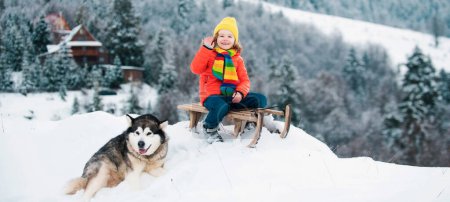 Photo for Boy with dog sledding on winter mountain, enjoying a sledge ride in a beautiful snowy winter nature. Winter fun kids activities - Royalty Free Image