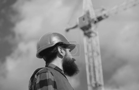 Foto de Male construction worker in work clothes and a construction helmet. Improvement and renovation. Brutal man builder. Engineer builder in uniform. Construction crane - Imagen libre de derechos