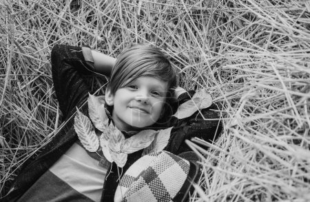Foto de Un niño sonriente jugando con hojas y mirando a la cámara. El niño yace en el heno. Lindo niño pequeño sosteniendo la hoja de oro en el fondo de la aldea granja - Imagen libre de derechos