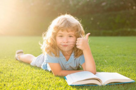 Téléchargez les photos : Joli garçon avec les pouces vers le haut livre de lecture sur l'herbe verte. Été plein air enfants éducation - en image libre de droit