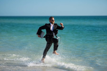 Téléchargez les photos : Homme d'affaires excité en combinaison humide courir en mer. Homme d'affaires drôle, concept d'affaires comique fou. Travail en ligne à distance. Des affaires d'été folles. Style de vie d'affaires amusant. Homme d'affaires freelance drôle - en image libre de droit