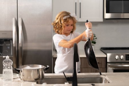 Foto de Divertidos chicos gemelos ayudando en la cocina con lavar los platos. Niños divirtiéndose con tareas domésticas. Niño lavando los platos en el fregadero de la cocina - Imagen libre de derechos