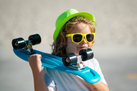 Foto de Feliz niño con estilo en gafas de sol y sombrero de verano de moda posando con monopatín al aire libre - Imagen libre de derechos