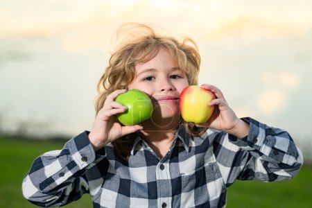 Téléchargez les photos : Mordre une pomme. Enfant tenant pomme, fond de parc d'été. Les enfants mangent de la pomme verte. Portrait de petit enfant souriant heureux avec des pommes. Aliments sains - en image libre de droit