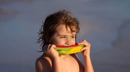 Téléchargez les photos : Le petit enfant mange de la pastèque rouge juteuse sur la plage. Vacances d'été pour enfants, fruits de saison et baies. Portrait extérieur d'enfant d'été. Face d'enfant d'été. Enfant à la pastèque, fruits d'été - en image libre de droit