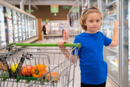Foto de Niño con carrito de compras en la tienda de comestibles. Niño comprando fruta en el supermercado. El niño compra verduras frescas en la tienda de comestibles. Niño eligiendo verduras. Alimento saludable - Imagen libre de derechos