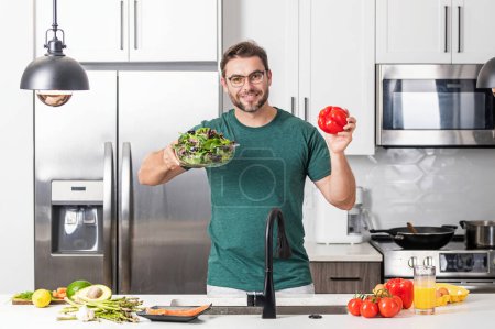 Photo for Taking care of health. Handsome man in casual t-shirt standing in the kitchen at home. Young man cooking vegetables in the kitchen. Dieting man with bowl of salad. Green salad healthy food concept - Royalty Free Image