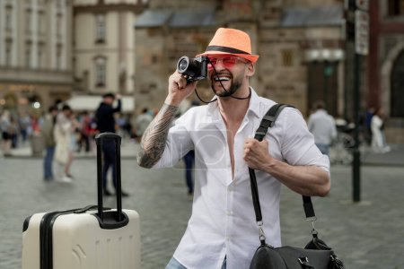 Téléchargez les photos : Voyageur homme avec lunettes de soleil, chapeau et appareil photo. Touriste homme à la mode chapeau jeans et lunettes de soleil avec valise marchant dans la rue. Homme avec bagages prêt à voyager en vacances - en image libre de droit