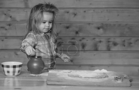 Téléchargez les photos : Cuisson bébé. Un gamin cuisine dans la cuisine. Petit garçon met la pâte dans un plat de cuisson en jouant avec de la farine - en image libre de droit