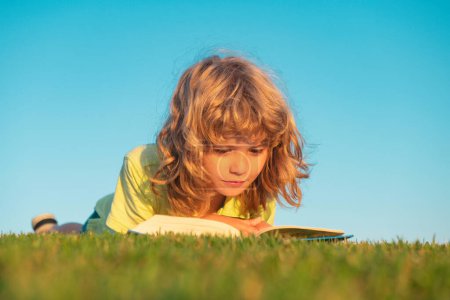 Photo for Kid read book in park. Child boy reading book laying on grass on grass and sky background with copy space outdoor - Royalty Free Image