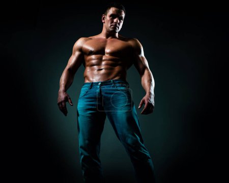 Photo for Sexy handsome tanned caucasian man in jeans, standing naked torso, showing six-pack abdominal abs, have perfect body, dark black background - Royalty Free Image