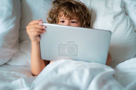 Foto de Concepto social y tecnológico. Internet para niños. Niño con tableta en la cama. Permiso parental, red en línea de seguridad. Niño jugando juegos en línea - Imagen libre de derechos