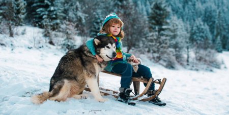 Téléchargez les photos : Un enfant en traîneau joue avec un chien dehors. Enfants d'hiver, vacances et Noël. Enfants étreinte chien husky - en image libre de droit