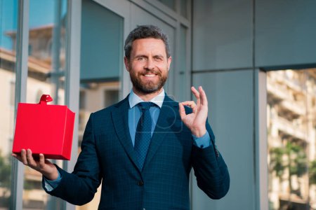 Téléchargez les photos : Portrait d'homme d'affaires avec ok signe en costume montrant boîte cadeau - en image libre de droit