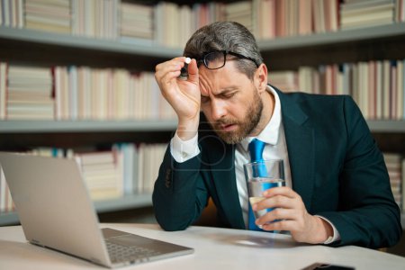 Téléchargez les photos : Millennial Homme d'affaires au bureau ayant mal à la tête. Homme d'affaires en costume travail sur ordinateur portable au bureau à domicile décoller des lunettes souffrent de migraine ou de maux de tête. Médicament pilule de migraine maux de tête - en image libre de droit