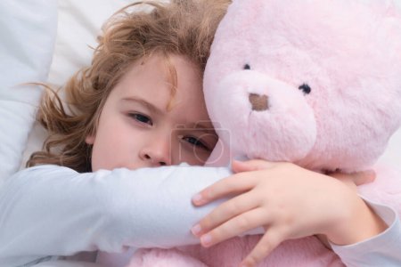 Photo for Child wakes up with toy teddy bear from sleep in bed. Kid wakes up in the morning in the bedroom. Favorite toy for sleep. Child dream. Dreamy kids face. Daydreamer child portrait close up - Royalty Free Image
