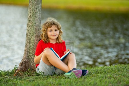 Foto de Lindo niño leyendo libro en el prado al aire libre. Escuela y educación. Naturaleza y parque. Aprendizaje temprano. Un niño leyendo un libro en el parque de verano. Concepto de recreación infantil sin artilugios - Imagen libre de derechos