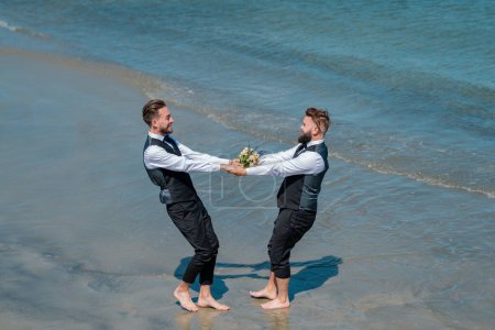 Photo for Gay grooms walking together on sea beach during Wedding day. Romantic men in sea water. Gay with partner with care and love. Gay couple holding bouquet of flower together during wedding ceremony - Royalty Free Image