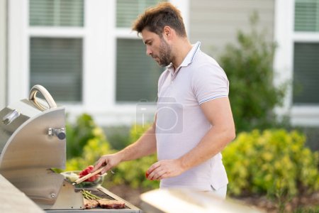 Photo for Man at a barbecue grill. Male cook preparing barbecue outdoors. Bbq meat, grill for picnic. Roasted beef. Cook preparing barbeque in the house yard. Barbecue and grill. Cook using barbecue tongs - Royalty Free Image