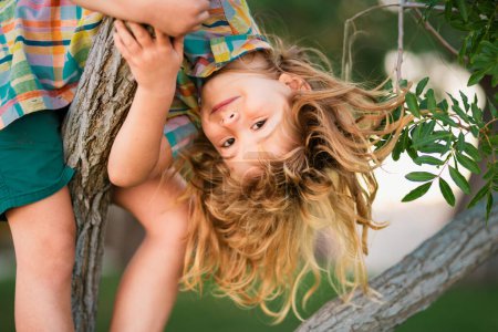 Foto de Niño rubio trepando al árbol. Feliz niño jugando en el jardín subiendo al árbol. Niño jugando y trepando a un árbol y colgando rama. Cara divertida de los niños - Imagen libre de derechos