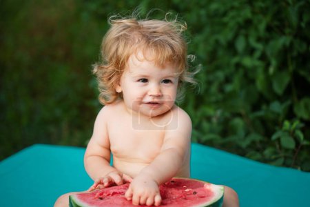 Téléchargez les photos : Petit bébé mangeant pastèque rouge dans le jardin assis sur l'herbe. Enfant mangeant de la pastèque dans le jardin. Les enfants mangent des fruits à l'extérieur. Mignon bébé jouant dans le jardin tenant une tranche de pastèque - en image libre de droit