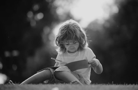Téléchargez les photos : Les enfants apprennent à distance. Elève préscolaire dans le parc sur herbe - en image libre de droit