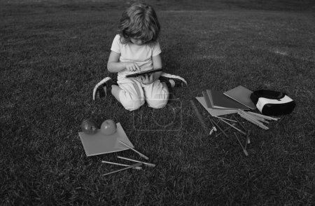 Foto de Los niños estudian afuera. Pequeño estudiante divertido con la tableta, sentarse en el césped en el parque, estudiando en el patio trasero de la escuela - Imagen libre de derechos