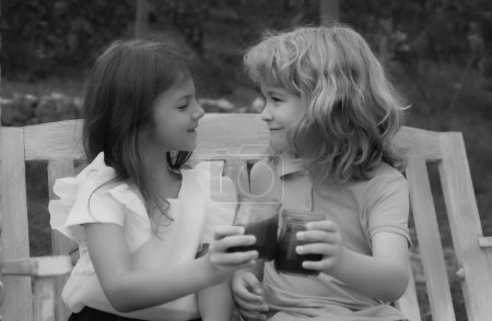 Foto de Dos niños beben batido, niños cóctel de verano al aire libre. Retrato de adorable hermano y hermana sonríen y ríen juntos mientras están sentados en columpio al aire libre. Feliz estilo de vida niños - Imagen libre de derechos