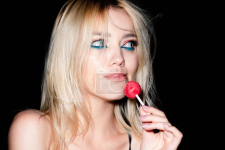 Photo for Portrait of sexy woman. Sensual blonde woman portrait with bright make up looking forward, licking red candy lollipop, studio black background - Royalty Free Image