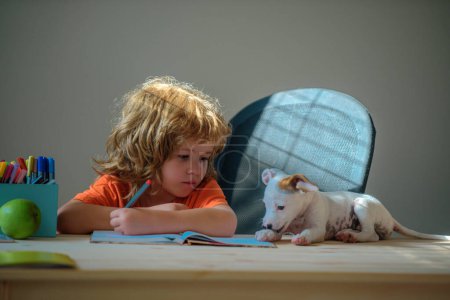 Foto de Lindo niño pequeño con cachorro perro haciendo su tarea. La educación de la infancia y el concepto de la escuela - lindo pequeño estudiante con la escritura de mascotas a cuaderno en casa - Imagen libre de derechos