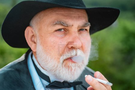Photo for Close up face portrait of handsome senior with gray beard smoking cigarette. Attractive elderly mature man. Senior glamour vintage man wearing suit and tie and hat. Gangster look, Mafia pimp - Royalty Free Image
