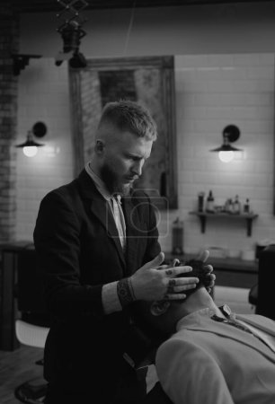 Téléchargez les photos : Un homme se fait couper les cheveux par un coiffeur. Un coiffeur en visite chez le coiffeur. Ciseaux de coiffeur et rasoir droit. Client barbu visitant le salon de coiffure - en image libre de droit