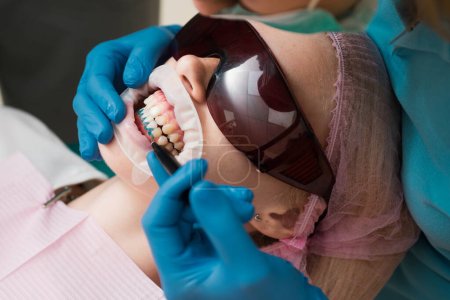 Téléchargez les photos : Dentiste guérir une patiente au cabinet dentaire dans un environnement agréable. Jeune femme blonde ouvrant la bouche alors qu'un dentiste non identifiable en gants de latex vérifie l'état de ses dents - en image libre de droit