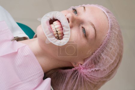Téléchargez les photos : Belle fille en chaise dentaire à l'examen chez le dentiste. Femme heureuse souriant sur un fond abstrait avec des lumières floues - en image libre de droit