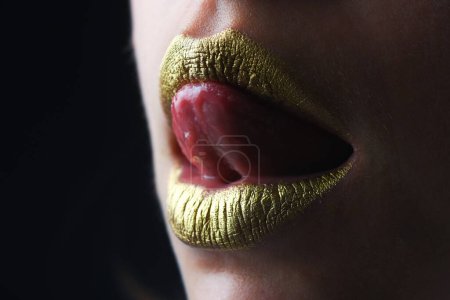 Téléchargez les photos : La langue sensuelle lèche les lèvres. Peinture dorée sur la bouche. Des lèvres dorées. Maquillage de luxe lèvres dorées. Lèvres dorées avec rouge à lèvres métallique créatif. Lèvre en métal doré. Femme sensuelle bouche, gros plan, macro - en image libre de droit
