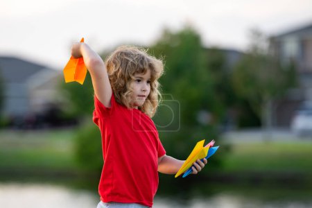 Foto de El niño sostiene aviones de papel. Avión de juguete en manos de niños al aire libre. Feliz chico inclinado y lanzando un avión de papel amarillo. Un chico jugando con un avión de papel. Niño jugando con avión de papel - Imagen libre de derechos