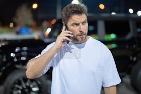 Photo for Angry man talking on phone outdoor. Angry hipster talking on phone near car on night city street. Serious angry guy talking on phone. Pensive handsome hipster look angry, talking on phone - Royalty Free Image