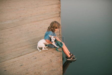 Foto de Niño pequeño con perro sentado en el parque cerca del agua - Imagen libre de derechos