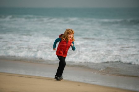Téléchargez les photos : Enfant traversant l'eau près du rivage le long de la plage de la mer. Un garçon court le long de la côte. Enfant coureur joggeur courir dans la nature. Mode de vie sain et actif des enfants - en image libre de droit