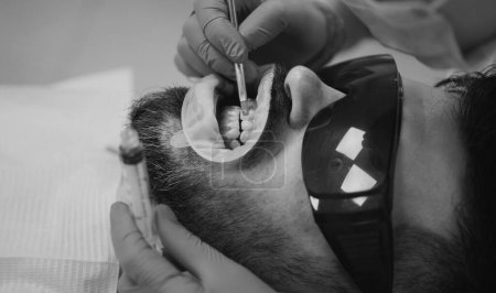 Téléchargez les photos : Dentiste féminine vérifiant les dents des patients avec miroir dans la clinique dentaire moderne. Patient ayant subi un examen dentaire à la lumière ultraviolette à la clinique dentaire. Jeune homme choisissant la couleur des dents chez le dentiste - en image libre de droit