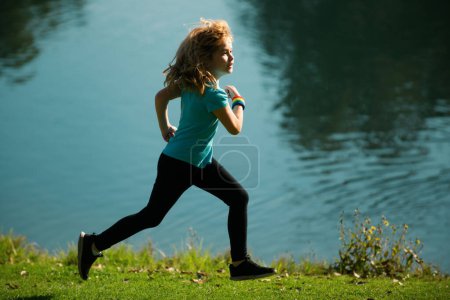 Téléchargez les photos : Enfant garçon courir ou faire du jogging près du lac sur l'herbe dans le parc. Garçon sportif coureur courir dans le parc d'été. Enfants actifs, enfants sportifs - en image libre de droit