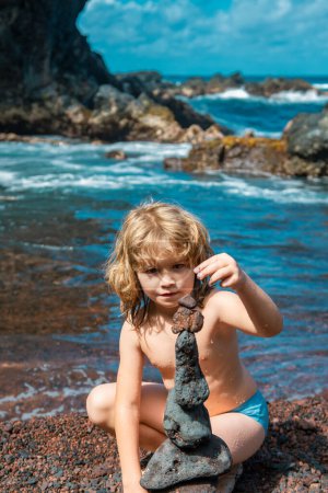 Téléchargez les photos : Garçon enfant faisant pile de cailloux pierre sur fond de mer pour spa, équilibre, méditation et thème zen - en image libre de droit