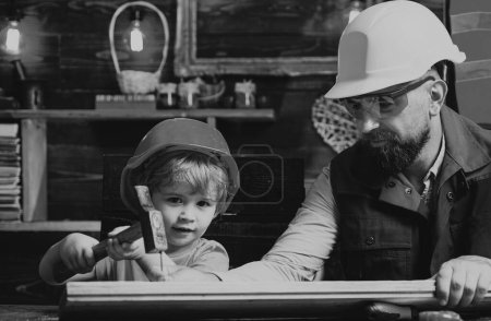 Foto de Lindo hijo y padre en sombrero duro sosteniendo tablón de madera y martillo. Chico ayudando a su padre en casa - Imagen libre de derechos