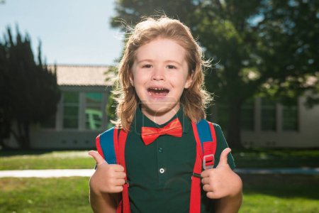 Téléchargez les photos : Joyeux petit écolier en uniforme scolaire avec un grand sac à dos debout près de l'école. Retour à l'école. Concept d'éducation préscolaire pour enfants - en image libre de droit